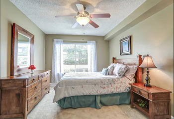Bedroom with wood ceiling fan and light in center of room.  Model bedroom with furniture and tan walls.  Window on back wall with white curtains.